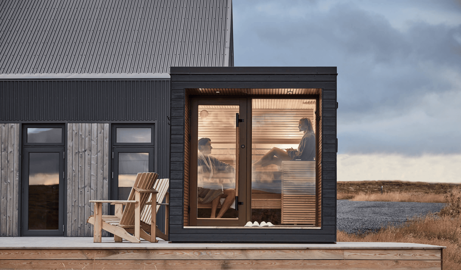 A compact and modern Auroom Arti outdoor sauna cabin with a large glass window, situated on a deck with Adirondack chairs in front, under a dark sky.