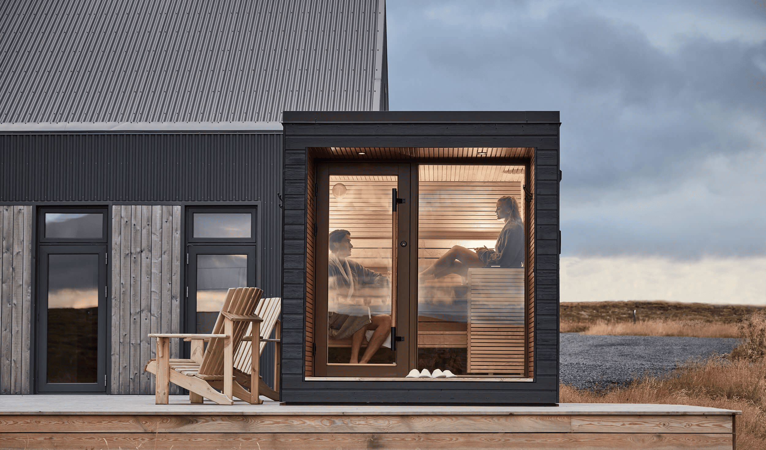 A compact and modern Auroom Arti outdoor sauna cabin with a large glass window, situated on a deck with Adirondack chairs in front, under a dark sky.